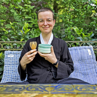 Eine Frau sitzt auf einem Sofa im Garten. In der Hand hält sie eine hellblaue Matchaschale und einen Matcha-Besen aus Bambus.