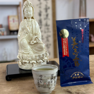 Eine buddhistische Statue, eine Teetasse gefüllt mir grünem Tee, eine Packung Sencha Monde Mishiro in blau mit goldenen Schriftzeichen