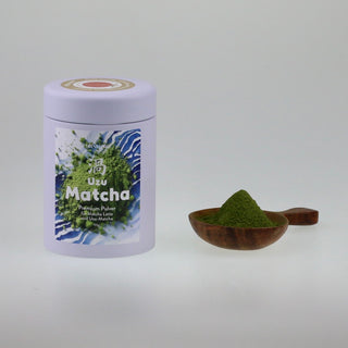 Frontale Ansicht einer hellvioletten Teedose mit der Aufschrift Uzu Matcha, daneben liegt ein kurzer, dunkler Holzlöffel gefüllt mit leuchtend grünem Matcha-Pulver.