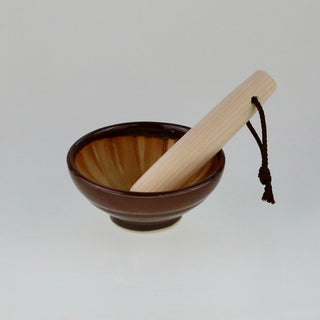 Schräge Aufsicht auf einen kleinen braunen Keramik-Mörser mit einem schräg hineingelegten hellen Holzstößel mit Kordel.