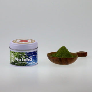 Frontale Ansicht einer hellen Teedose mit blau-weißem Etikett und der Aufschrift „Uzu Matcha“, daneben liegt ein dunkler Holzlöffel gefüllt mit leuchtend grünem Matcha-Pulver.