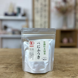 Eine silbrige Packung Benifuuki-Pulver steht auf einem alten Holztisch, im Hintergrund sieht man eine japanische Kalligraphie