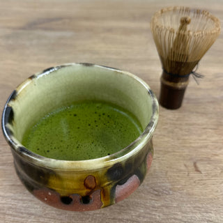 Matchaschale Bidoro in orange, grün und schwarz, gefüllt mit schaumigem Matcha, auf einem Holztisch. Daneben steht ein Matchabesen