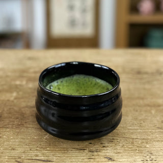 Die schwarz glasierte Matchaschale Kuroi, gefüllt mit aufgeschäumtem Matcha, steht auf einem antiken Holztisch