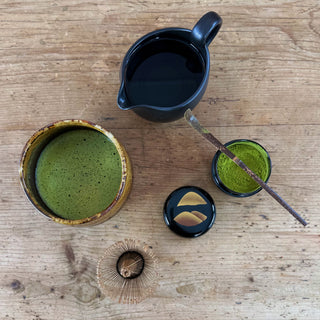 Mehrere Objekte stehen auf einem hellen Holztisch und wurden von oben fotografiert. Links die Matchaschale Tora mit bernsteinfarbener Glasur, gefüllt mit grünem Matcha. Oben im Bild ein mit Wasser gefülltes schwarzes Abkühlgefäß. Rechts im Bild eine mit Matchapulver gefüllte Matchadose, über der ein dunkelbrauner Matchalöffel liegt. Daneben der schwarze Dckel dieser Dose mit einem goldenen Motiv darauf und ein Matchabesen.