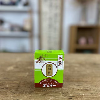 Eine grüne Matcha-Packung mit der Aufschrift "Chidori no Shiro". Oben links befindet sich eine Zeichnung eines traditionellen japanischen Schreins. Die Verpackung ist mit japanischen Schriftzeichen versehen und hat eine braunrote Basis. Im Hintergrund sind unscharf Regale und Dekorationsgegenstände zu erkennen, die eine gemütliche Raumatmosphäre andeuten.