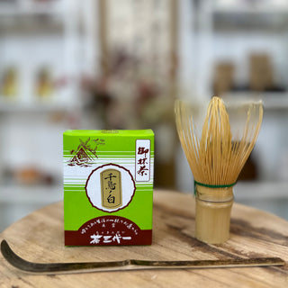 Verpackung von Shinkoko Matcha Chidori no Shiro in einer grünen Schachtel, daneben ein Matchabesen namens Chasen shin natur und ein Matchalöffel namens Chashaku schwarz auf einem Holztisch, im Hintergrund ein unscharfer japanischer Dekor.