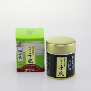 Links ist die grüne Verpackung des Matcha "Chitose" mit einer Zeichnung eines japanischen Schreins und japanischen Schriftzeichen auf braunrotem Hintergrund. Rechts daneben steht die Matcha-Dose mit schwarzem Etikett und goldenem Deckel. Der Hintergrund ist schlicht und weiß.