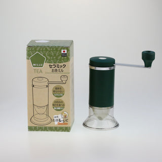 Schräge Aufsicht auf eine stehende, dunkelgrüne Handmühle für Tee mit Kurbel neben ihrer bedruckten Originalverpackung.