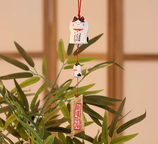 Furin Keramikglocke „Maneki Neko Kuroshiro“ als Windspiel, hängend vor grünen Blättern dekoriert.