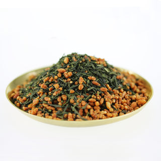 Eine große Portion Genmaicha ist auf einem goldenen Tellerchen vor einem weißen Hintergrund aufgetürmt. Der Genmaicha setzt sich zusammen aus dunkelgrünen, länglich gerollten Teeblättern und goldgelb gerösteten Maiskörnern, ungefähr im Verhältnis 50:50.