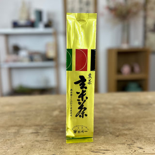 Eine längliche Packung Genmaicha in goldener Farbe steht auf einem hellen Holztisch