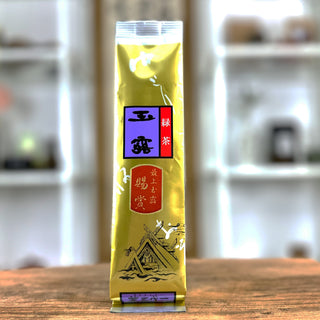 Eine goldene Packung Gyokuro Soshou, mit Etiketten in den Farben rot, lila und orange, auf denen japanische Schriftzeichen sind, sowie der Zeichnung eines japanischen Schreins. Die Packung steht auf einem antiken Holztisch.