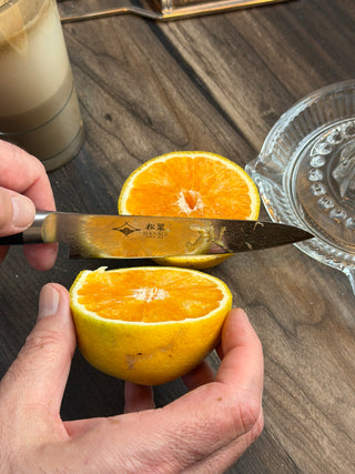 Eine Hand schneidet mit dem japanischen Petty-Messer Ranmaru eine Orange auf einem Holztisch, daneben stehen eine gläserne Zitruspresse und ein Glas mit einem hellen Getränk.