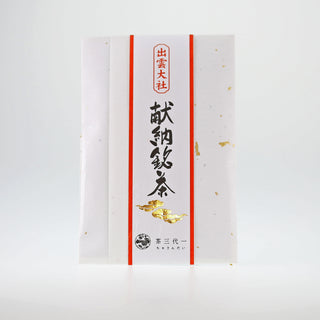 Eine Packung "Izumo Taisha Sencha" vor einem weißen Hintergrund. Die Verpackung hat im Zentrum eine japanische Kalliegraphie, verziert mit goldenen Wolken. Sie wird links und rechts von grellroten Streifen umrahmt. Die Packung selbst ist aus schwerem, weißem Papier. 