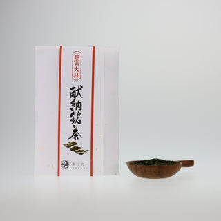 Eine weiße Packung Izumo Tencha Sencha, mit großen japanischen Schriftzeichen und roten Balken verziert, steht vor einer weißen Leinwand. Daneben befindet sich in einem braunen, runden Holzgefäß ein Häufchen der dunkelgrünen Teeblätter.