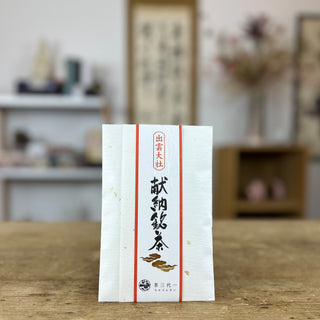 Eine Packung "Izumo Taisha Sencha" in weiß, mit einer schwarzen Kalligraphie und goldenen Wolken, umrahmt von zwei roten Streifen, steht auf einem Holztisch. im Hintergrund kann man vage einen Blumenstrauß, ein Wandbild mit Schriftzeichen und zwei Regale erkennen.