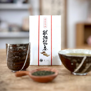 Vier Gegenstände stehen auf einer Holzplatte. Im Hintergrund eine Packung "Izumo Taisha Sencha". Sie ist weiß, mit zwei senkrechten roten Streifen, zwischen denen eine japanische Kalligraphie und zwei goldene Wolken zu sehen sind. Im Vordergrund sieht man ein Holztellerchen mit einem Häufchen Teeblättern. Links steht die Teetasse "Hikari", rechts die Schale "Hikari". Sie sind dunkelbraun, mit weißen diagonalen Streifen verziert.