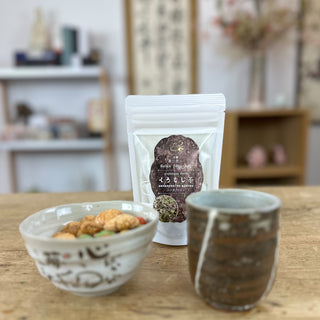 Links im Bild steht unsere Schale "Neko". Sie ist grau, mit einem Katzenmotiv darauf, und gefüllt mit bunten japanischen Snacks. Im Hintergrund steht die weiße Packung Kuromoji-Cha. Rechts steht unsere Teetasse "Hikari" in Dunkelbraun mit weißen , diagonalen Streifen.