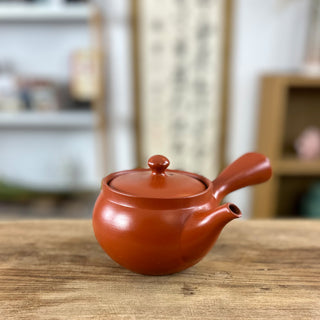 Eine braune Kyusu-Teekanne mit einem kurzen Ausguss und einem runden Deckel, präsentiert auf einem Holztisch. Im Hintergrund sind unscharf Regale und Deko-Objekte zu sehen.