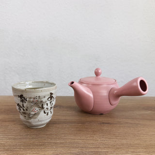 Eine rosafarbene Kyusu-Teekanne namens "Kyusu Ichigo" aus Keramik. Daneben steht eine graue Tasse namens "Usagi-Teetasse" mit japanischen Schriftzeichen und Hasenmotiven. Beide stehen auf einer Holzoberfläche vor einer weißen Wand.