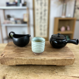 Die schwarze Kyusu-Teekanne "Kage" aus Keramik mit rundem Deckel und seitlichem Ausguss steht auf einem Holztisch. Daneben stehen die altweiße, gerippte Teetasse "Haru" mit grünem Tee und das schwarze Abkühlgefäß "Yuzamashi".