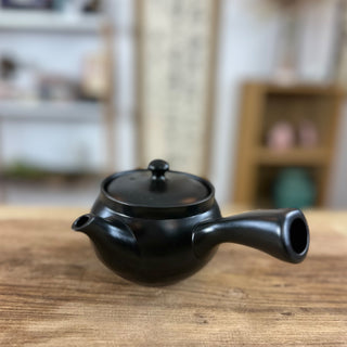 Die schwarze Kyusu-Teekanne "Kage" aus Keramik mit rundem Deckel und seitlichem Ausguss. Sie steht auf einem Holztisch. Im Hintergrund sind unscharfe Regale und Dekorationsgegenstände, die eine gemütliche Raumatmosphäre andeuten.