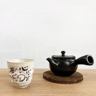 Die schwarze Kyusu-Teekanne "Kage" aus Keramik mit rundem Deckel und seitlichem Ausguss steht auf einem Holzuntersetzer. Daneben steht die graue Teetasse "Neko" mit japanischen Schriftzeichen und niedlichen Katzenmotiven. Beide Objekte stehen auf einer Holzoberfläche vor einer weißen Wand.