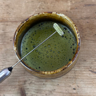 Ein elektrischer Matcha-Aufschäumer aus Edelstahl liegt quer über einer Tasse fertig aufgeschäumtem Matcha. Beide Objekte sind aus der Vogelperspektive aufgenommen und stehen auf einem antiken Holztisch.