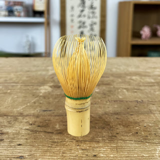 Traditioneller Matchabesen namens 'Chasen Shin natur' aus naturfarbenem Bambus mit feinen Borsten und grüner Schnur, auf einem Holztisch. Im Hintergrund sind ein Regal, eine japanische Schriftrolle und Dekorationsgegenstände zu sehen.