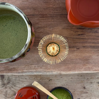 Auf einem Holztisch liegen verschiedene Teezubehörteile: eine grün-schwarze Matchaschale namens 'Bidoro' mit aufgeschäumtem Matcha-Tee, ein rotes Abkühlgefäß, ein traditioneller Bambusbesen namens 'Chasen Shin natur' und eine Matcha-Teedose namens 'Negoro' mit rot-schwarzem Muster, gefüllt mit grünem Matcha-Pulver. Ein Bambus-Teelöffel liegt auf der geöffneten Dose. Die Szene ist von oben aufgenommen.