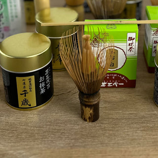 "Verschiedene Teezubehörteile und Produkte stehen auf einem Holztisch: ein traditioneller Matchabesen namens 'Chasen Shin schwarz', Teedosen mit goldenen Deckeln und eine grüne Verpackung mit dem Matcha 'Chidori no Shiro' aus dem Sortiment. Ein Bambus-Teelöffel ruht auf der Verpackung.