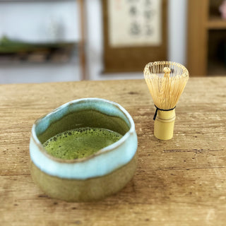 Auf einem Holztisch stehen eine Matchaschale namens 'Sora' mit grünem Matcha-Tee und ein Matchabesen 'Chasen basic natur'. Im Hintergrund sind ein Regal, eine japanische Schriftrolle und Dekorationsgegenstände zu sehen.