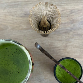 Drei Gegenstände werden in der Vogelperspektive gezeigt. Oben im Bild sieht man einen Matchabesen. Unten rechts ist eine mit Matcha gefüllte Matchaschale "Bidoro" mit oranger und grüner Glasur. Rechts unten ist eine mit Matchapulver gefüllte Matchadose in schwarz, mit einem dunkelbraunen Matchalöffel quer über der geöffneten Dose.