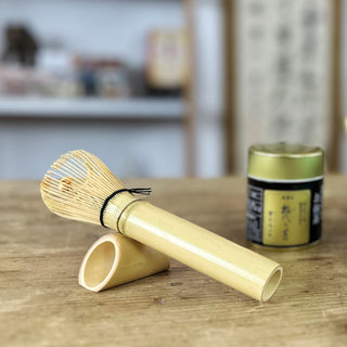 Traditioneller Matchabesen namens 'Chasen Tenmoku natur' aus naturfarbenem Bambus mit feinen Borsten, liegend auf einem passenden Bambus-Halter auf einem Holztisch. Im Hintergrund steht eine Teedose mit goldenem Deckel. Ein Regal und eine japanische Schriftrolle sind ebenfalls zu erkennen.