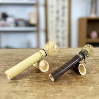 Zwei traditionelle Matchabesens namens 'Chasen Tenmoku' in den Farben 'natur' und 'schwarz', liegend auf passenden Bambus-Haltern auf einem Holztisch. Im Hintergrund sind ein Regal und eine japanische Schriftrolle zu sehen.