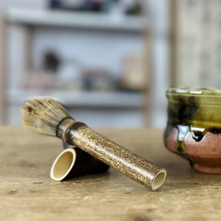 Traditioneller Matchabesen namens 'Chasen Tenmoku schwarz' aus dunklem Bambus mit feinen Borsten, liegend auf einem passenden Bambus-Halter auf einem Holztisch. Im Hintergrund steht eine grün-schwarze Matchaschale namens 'Bidoro'. Ein Regal und eine japanische Schriftrolle sind ebenfalls zu erkennen.