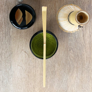 Ein traditioneller Bambus-Matchalöffel namens "Chashaku natur" liegt quer über einer geöffneten schwarzen Matcha-Teedose, die mit grünem Matcha-Pulver gefüllt ist. Der Deckel der Dose liegt daneben auf einem Holztisch. Unten im Bild steht ein Bambus-Matchabesen (Chasen) auf einem weißen Keramikhalter.