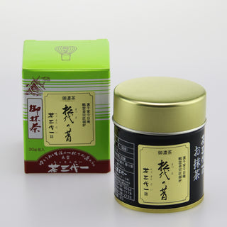 Links ist die leuchtend grüne Verpackung des Matcha "Matsuyo no Mukashi" mit einer Zeichnung eines japanischen Schreins und japanischen Schriftzeichen auf braunrotem Hintergrund zu sehen. Rechts daneben steht die dazugehörige Matcha-Dose mit schwarzem Etikett und goldenem Deckel. Der Hintergrund ist schlicht und weiß.