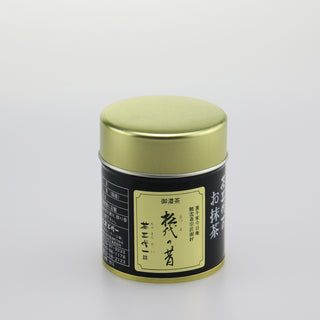 Eine Matcha-Dose "Matsuyo no Mukashi" mit schwarzem Etikett und goldenem Deckel. Die Dose zeigt japanische Schriftzeichen auf einem hellen Etikett. Der Hintergrund ist neutral und weiß.