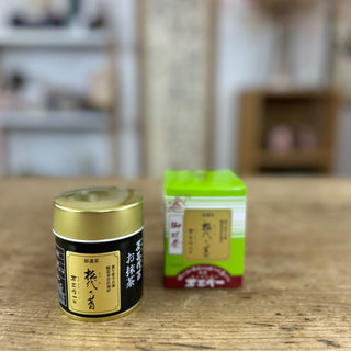 Im Vordergrund steht die Matcha-Dose "Matsuyo no Mukashi" mit schwarzem Etikett und goldenem Deckel. Dahinter befindet sich die leuchtend grüne Verpackung mit einer Zeichnung eines japanischen Schreins und japanischen Schriftzeichen auf braunrotem Hintergrund. Der Hintergrund zeigt unscharfe Regale und Dekorationsgegenstände, die eine gemütliche Raumatmosphäre andeuten.