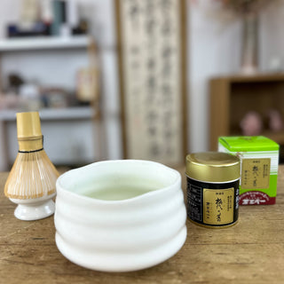  Im Vordergrund stehen die Matcha-Dose "Matsuyo no Mukashi" mit goldenem Deckel und die weiße Keramik-Matchaschale "Shiroi". Links ist ein traditioneller Matchabesen zu sehen, im Hintergrund die grüne Verpackung des Matcha und unscharfe Regale sowie Dekorationsgegenstände, die eine gemütliche Atmosphäre schaffen.