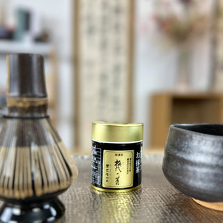  Im Vordergrund steht die Matcha-Dose "Matsuyo no Mukashi" mit schwarzem Etikett und goldenem Deckel. Daneben sind ein traditioneller Matchabesen und die dunkle Keramik-Matchaschale "Yoru". Der Hintergrund zeigt unscharfe Regale und Dekorationsgegenstände, die eine gemütliche Atmosphäre schaffen.