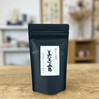 Eine schwarze Packung "Monde Mishiro Sencha" steht auf einem hellen Holztisch. Auf der Vorderseite der Packung sieht man auf einem weißen Etikett japanische Schrift in den Farben Schwarz und Rot.