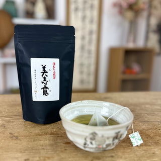 Links im Bild steht eine Packung "Monde Mishiro Sencha". Sie ist schwarz und hat auf der Vorderseite ein weißes Etikett, auf dem Schriftzeichen angebracht sind. Daneben steht die hellgrüne "Jizo"-Schale, in der sich grüner Tee und ein Teebeutel befinden.