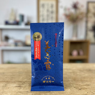 Eine blaue Packung "Monde Mishiro Sencha", auf der goldene und rote Schriftzeichen sowie ein hellrotes Banner und ein goldener Aufkleber prangen, steht auf einer alten Holztischplatte. Im Hintergrund kann man an einer Wand ein Rollbild mit japanischen Schriftzeichen erkennen, sowie einen Blumenstrauß und Regale mit Dekorationsobjekten.