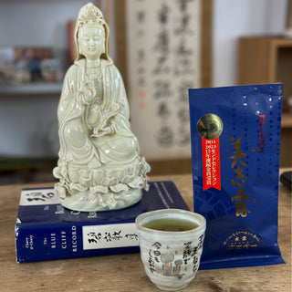 Vier Gegenstände sind auf einem Holztisch arrangiert. Im Vordergrund steht eine "Jizo"-Teetasse in hellgrau, gefüllt mit grünem Tee. Rechts daneben ist eine blaue Packung "Monde Mishiro Sencha" mit goldenen Schriftzeichen darauf zu sehen. Links davon liegt ein blaues Buch, auf dem eine buddhistische Statue aus weißer Keramik sitzt. Die Figur stellt ein weibliches Wesen dar. 