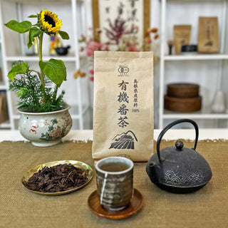 Verpackung von Shinkoko Bio-Bancha geröstetem Tee in braunem Papierbeutel, daneben eine Schale mit losen Teeblättern, eine traditionelle japanische Teekanne und eine Teetasse namens Hikari, im Hintergrund eine Sonnenblume in einer Vase.