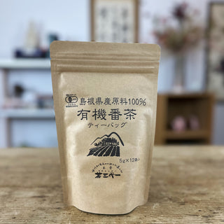 Verpackung von Shinkoko Bio-Bancha geröstetem Tee in Teebeuteln auf einem Holztisch, im Hintergrund ein unscharfes japanisches Dekor.