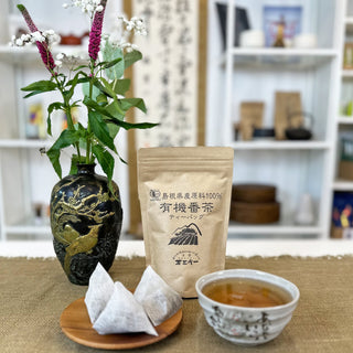 Verpackung von Shinkoko Bio-Bancha geröstetem Tee in Teebeuteln, daneben zwei dreieckige Teebeutel auf einer Holzschale und eine Teeschale namens Usagi mit Tee, im Hintergrund eine dekorative Vase mit Blumen und japanischem Dekor.